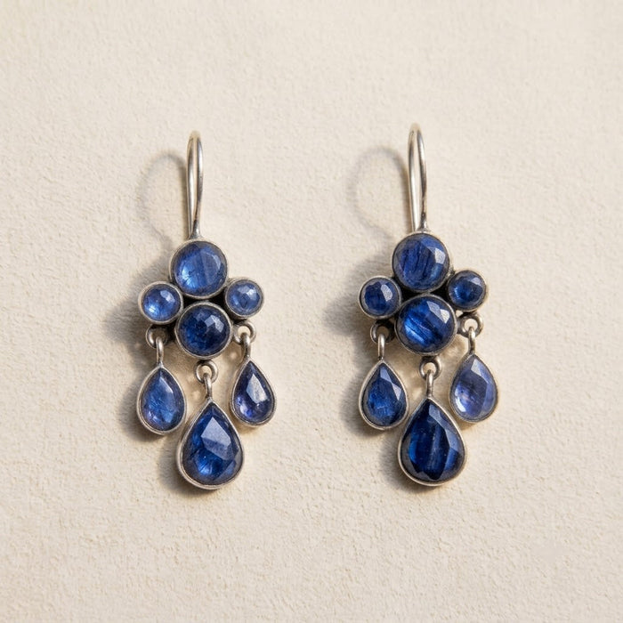 A pair of silver drop earrings featuring deep blue teardrop and round gemstones, displayed on a textured beige background.