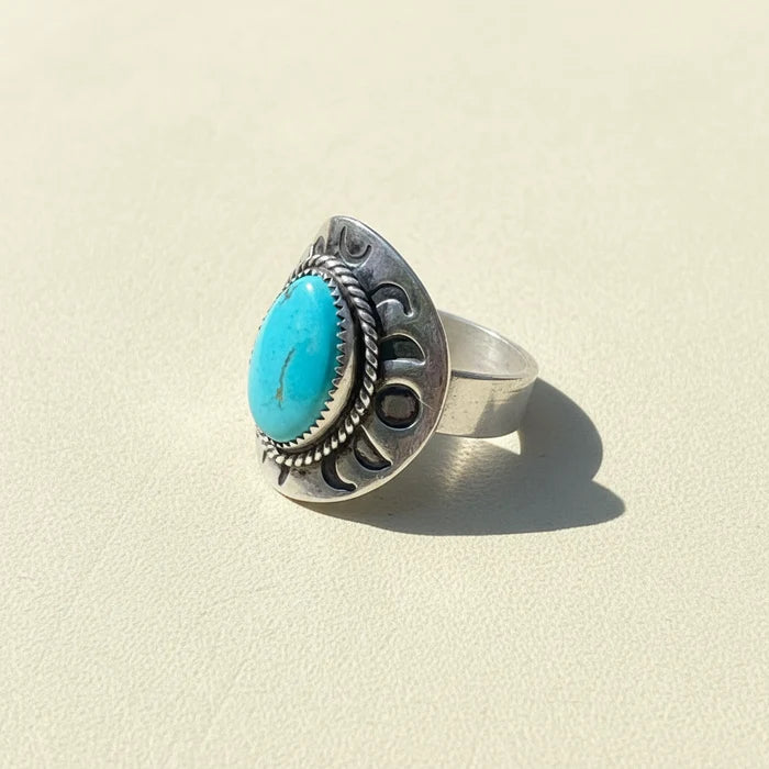 A sterling silver ring with a large oval turquoise cabochon and decorative engraved details, resting on a weathered wooden surface outdoors.